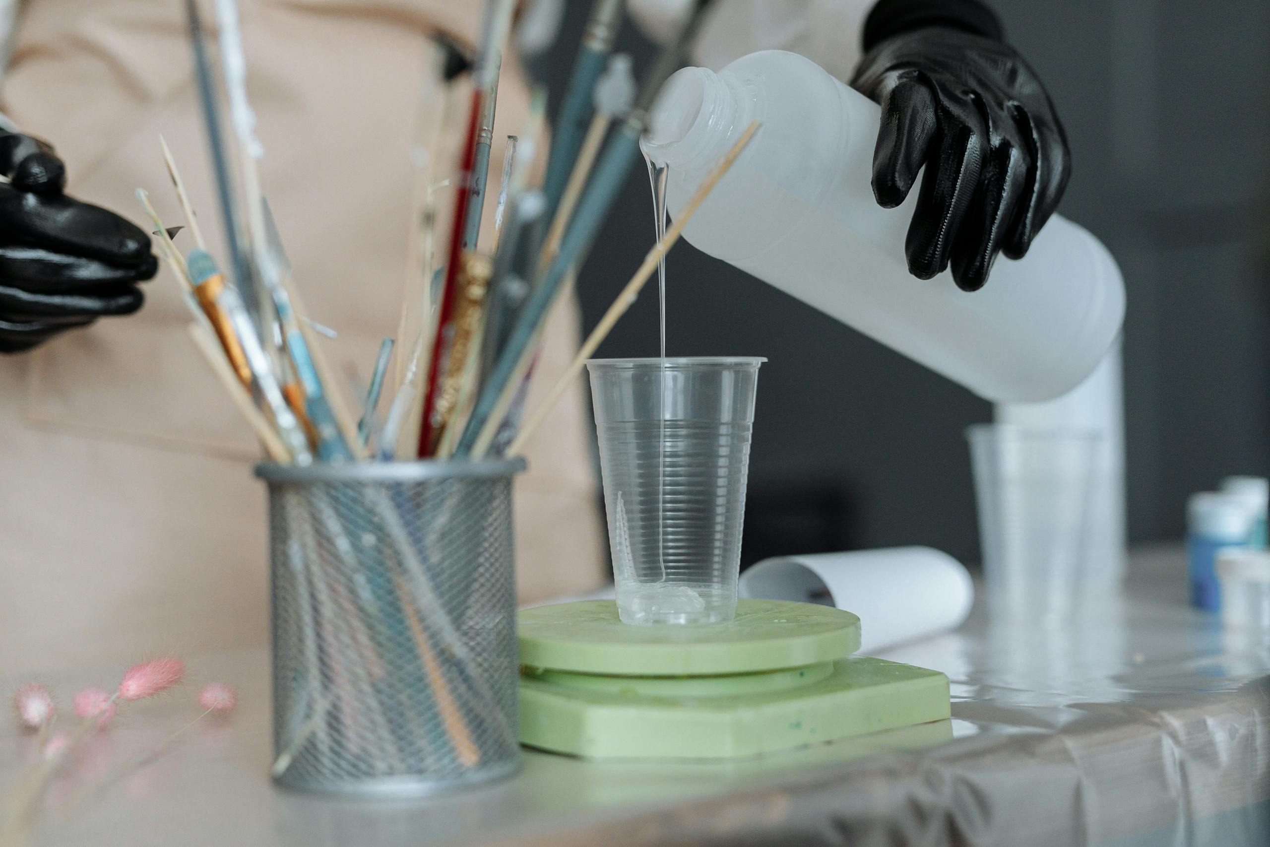 Close-up of resin being poured into a cup amidst art tools and safety gloves.