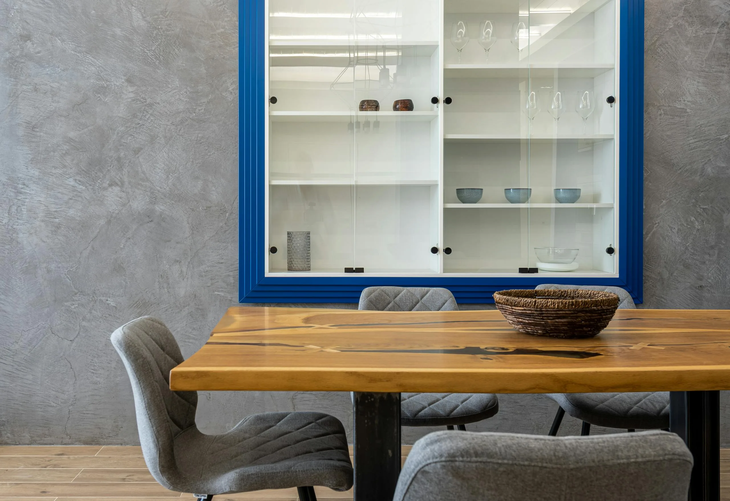 Comfortable chairs at wooden dinner table with bowl placed near gray wall with glass cabinet with glassware and dishes in modern kitchen